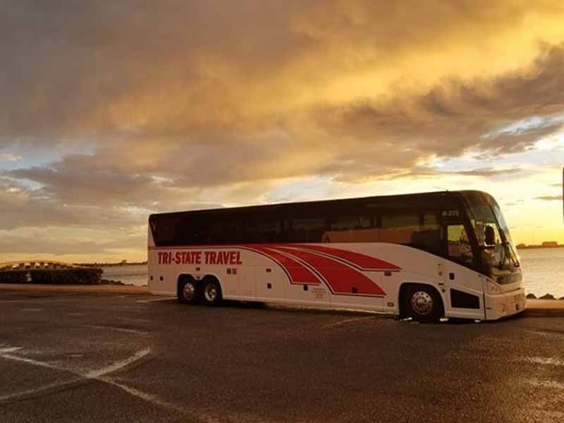 White tour bus with red graphics parked on an open lot at sunset under a cloudy sky