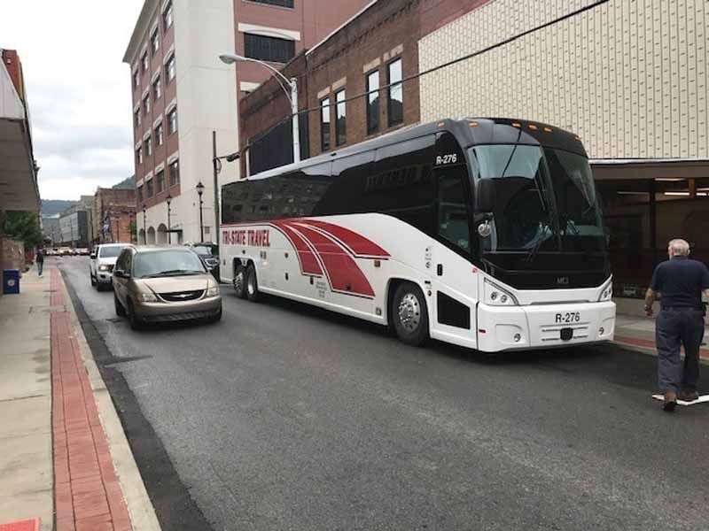 White coach bus parked on a city street beside a silver car and a pedestrian near brick buildings.