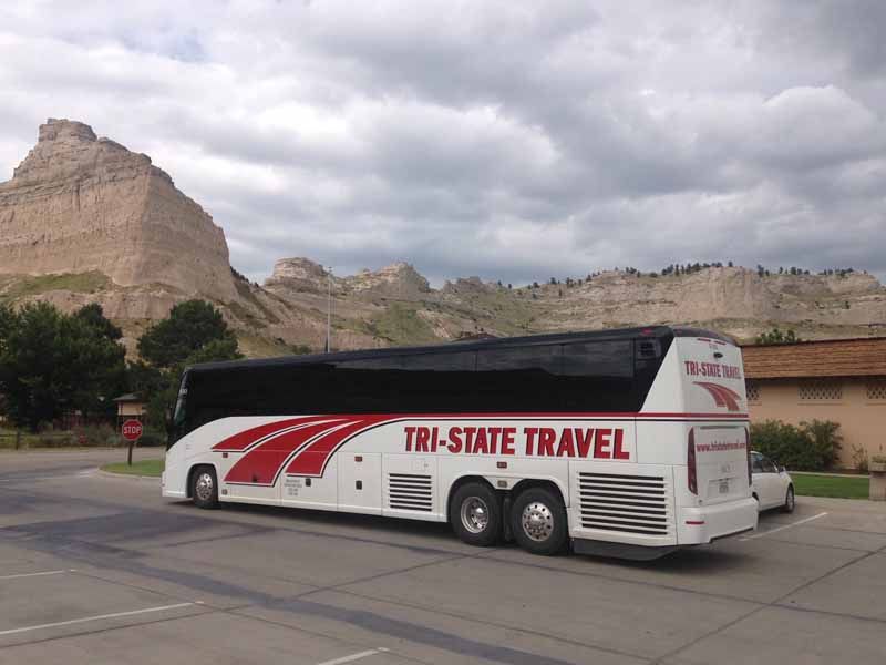 White Tri-State Travel bus parked beside rocky mountains under cloudy skies