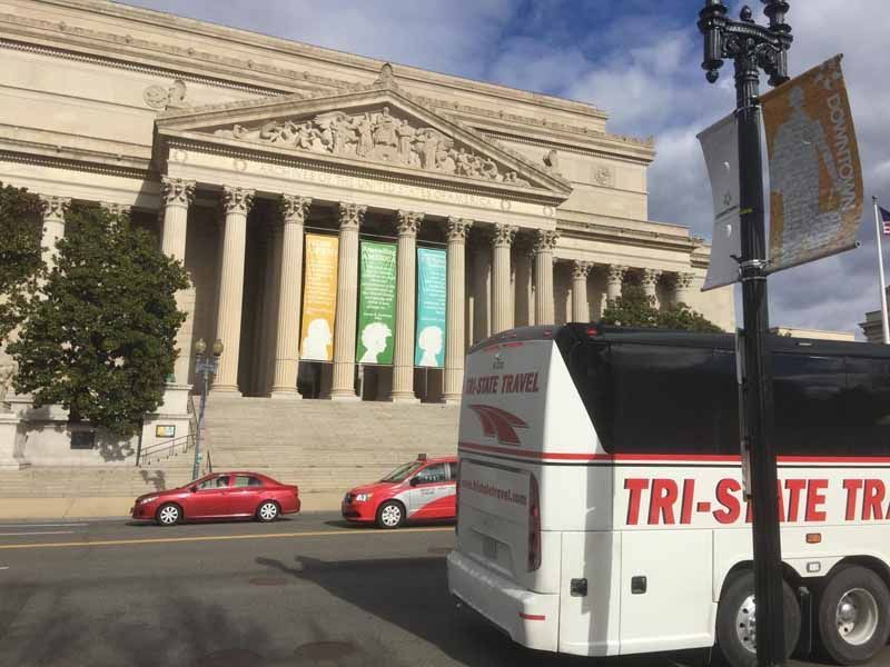 Classical museum facade with banners; red cars and a Tri-State bus passing in front