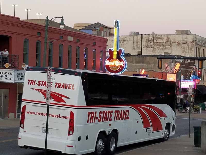 White Tri-State Travel tour bus parked on a city street near a neon guitar sign at dusk