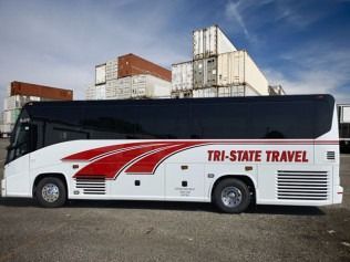 White Tri-State Travel coach bus parked outdoors in front of stacked shipping containers