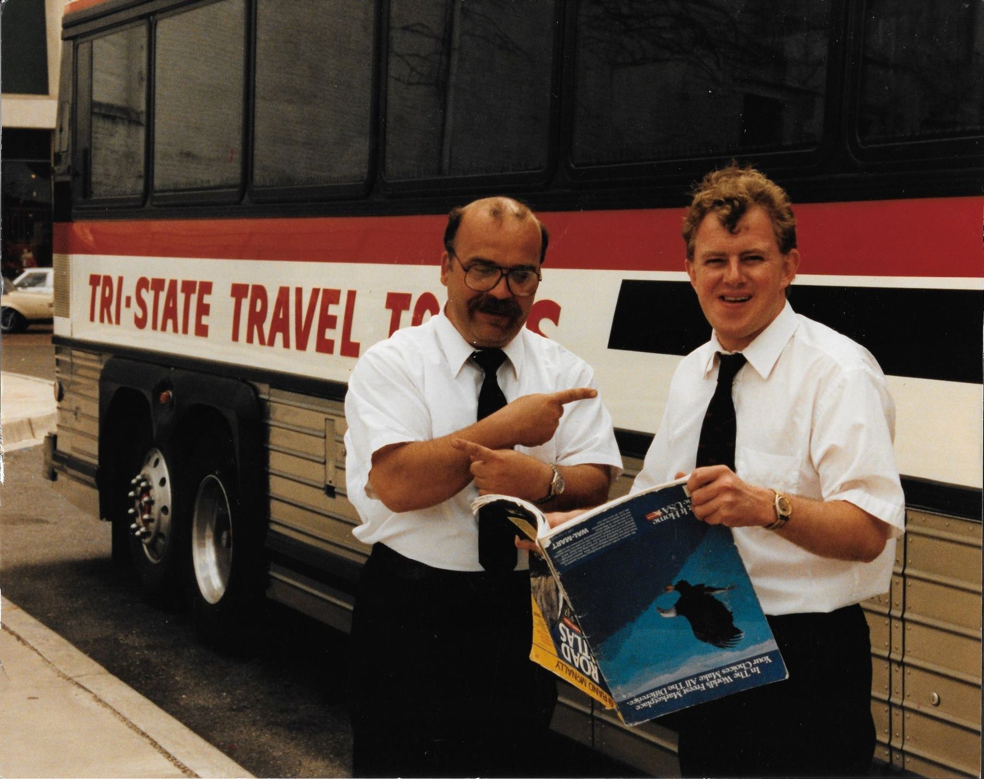 Two men standing by a bus, pointing at a brochure outside a travel office