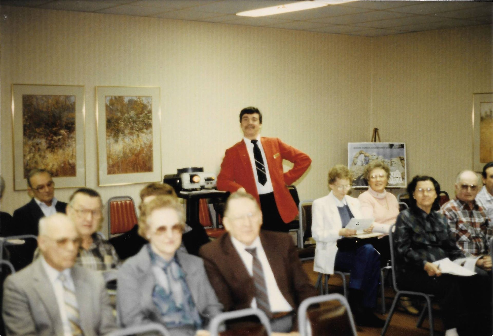 People seated in a community room while a man in a red jacket stands at the back.