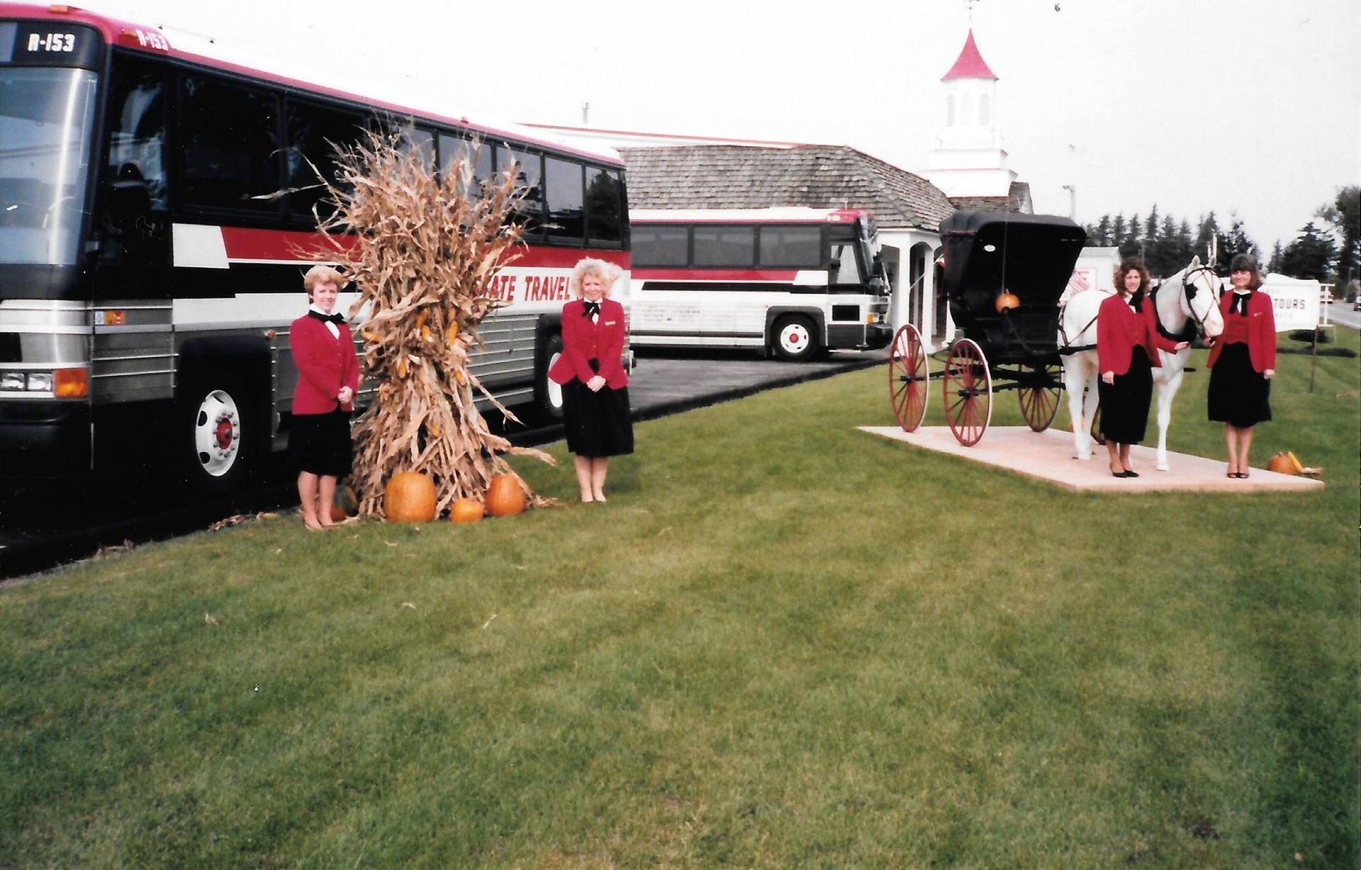 People on a lawn beside two buses, with fall decorations and a cloudy sky.