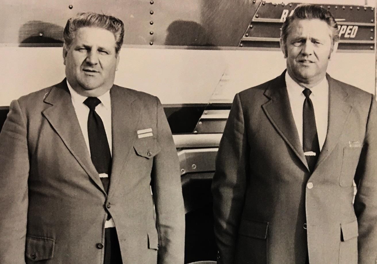 Two men in suits standing in front of a vehicle or trailer