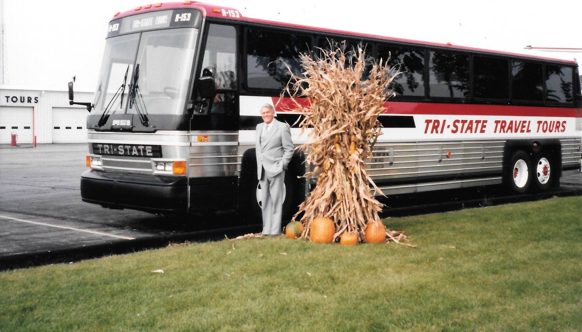 Gray-and-red bus beside a scarecrow and pumpkins on a grassy lawn