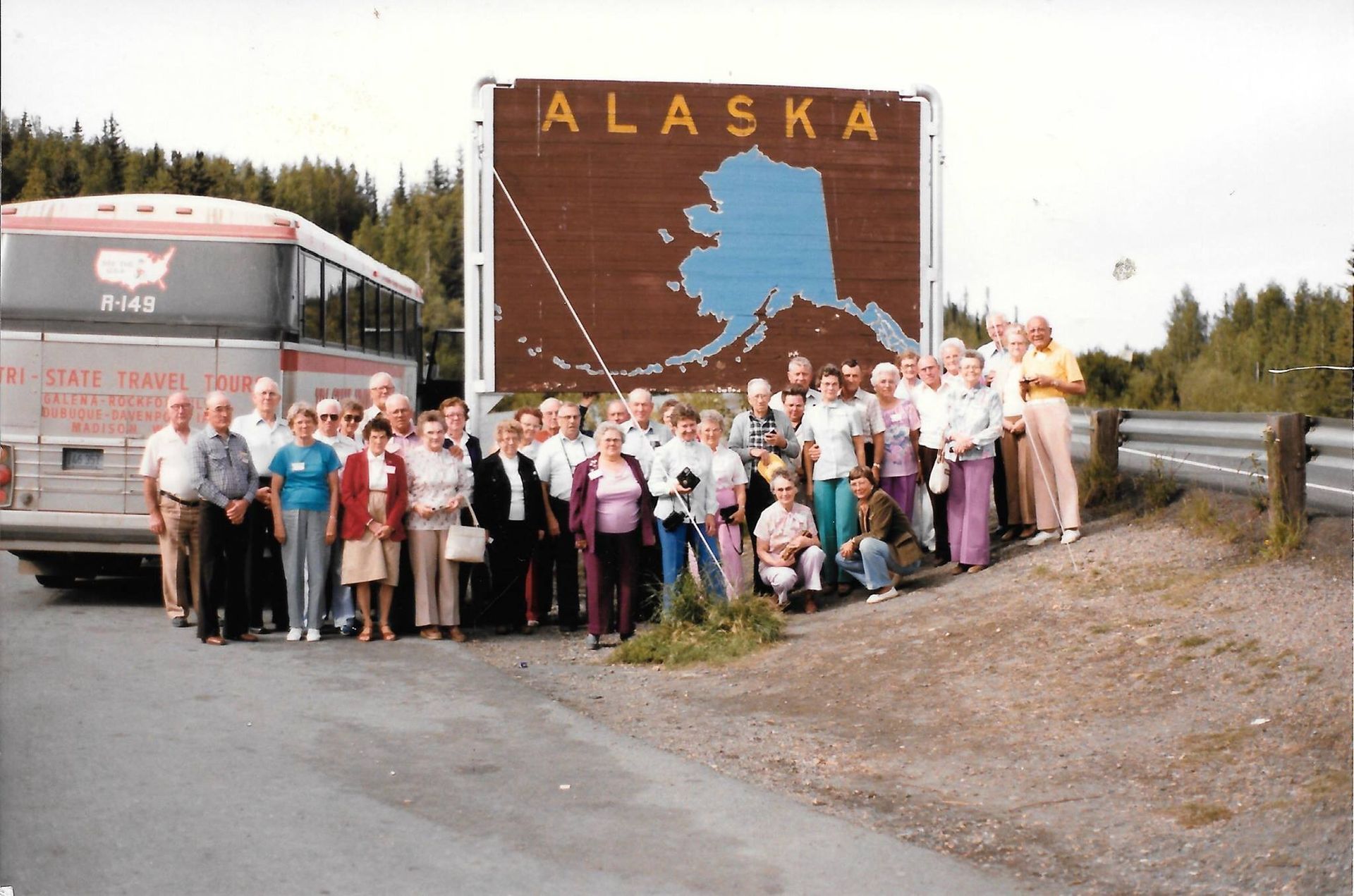 Group posing by an Alaska roadside sign beside a bus and guardrail in a rural area