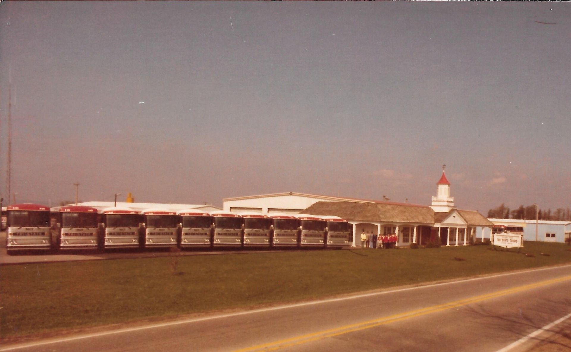Long row of buses parked by a roadside beside a white building with a red-roofed tower under a clear sky