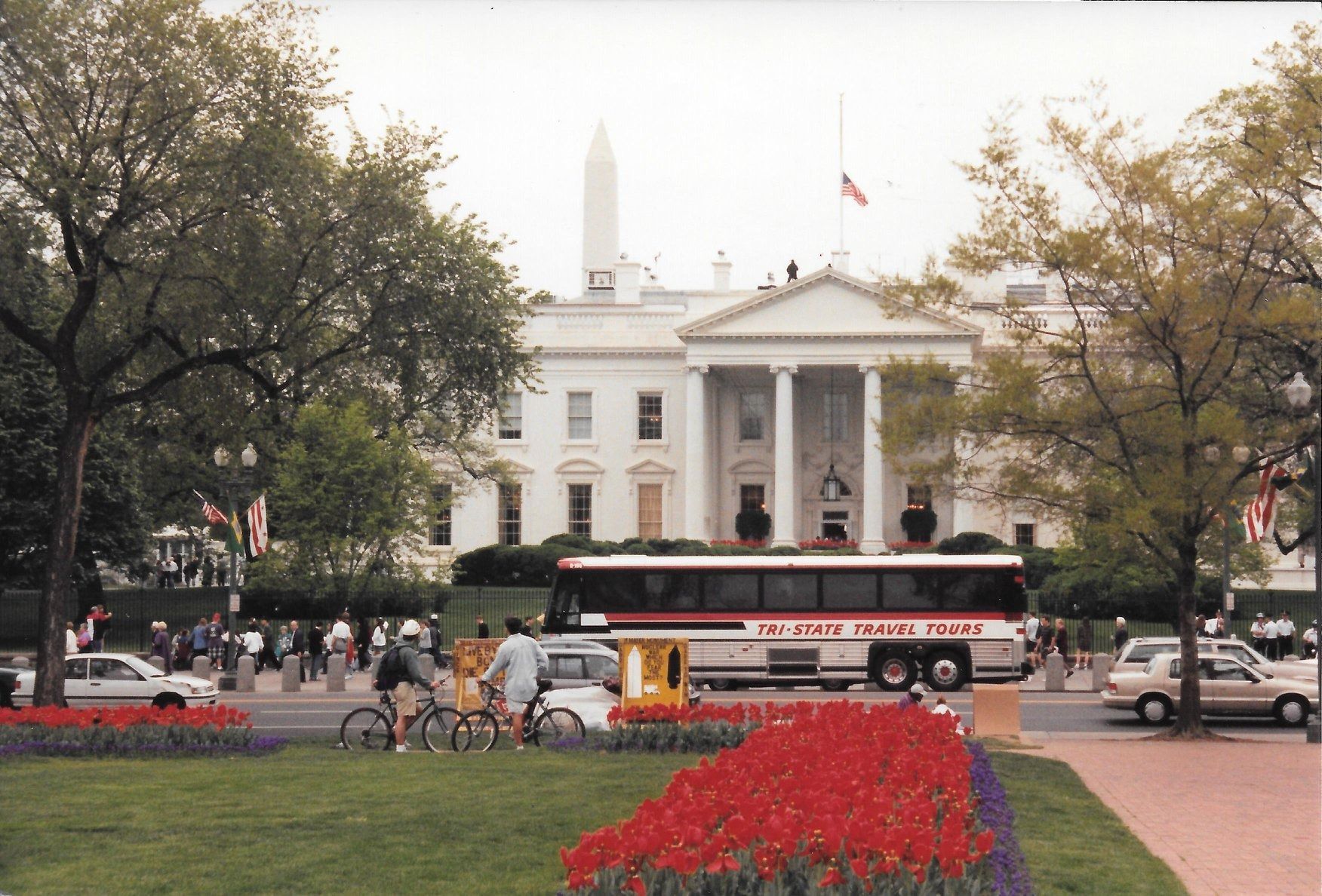 White House with a bus and people in front, framed by trees and red flowers in the foreground