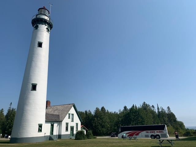 Tall white lighthouse beside a small house, with a tour bus parked on a grassy coast under a clear blue sky