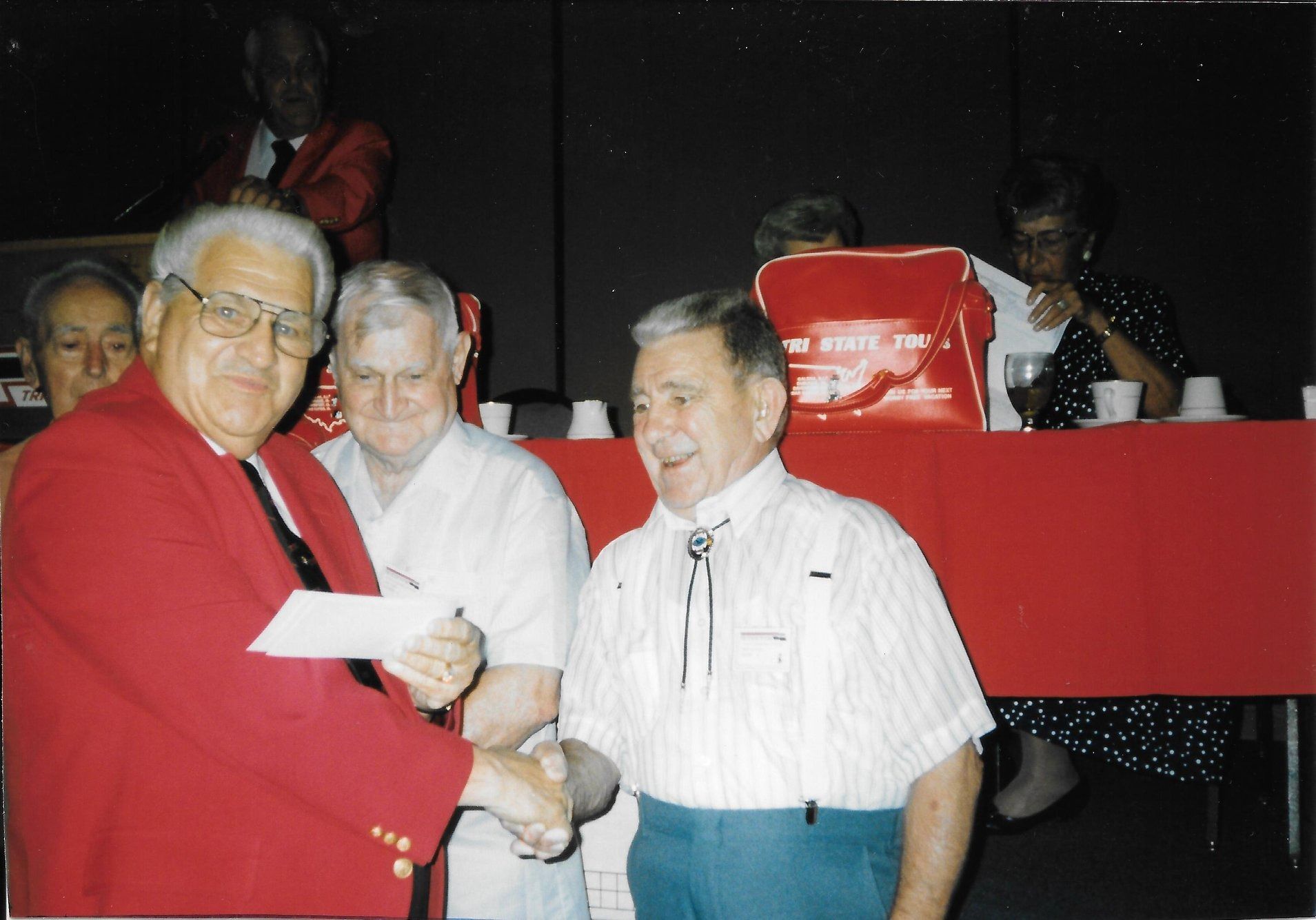 Three men stand at a banquet table, smiling and shaking hands in front of a red backdrop.