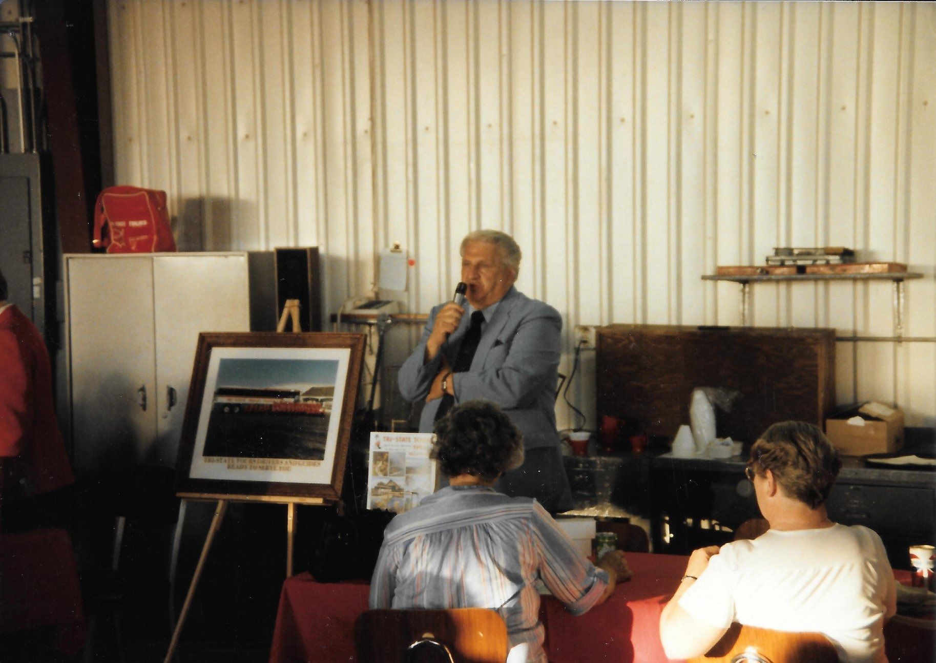 Man speaking into a microphone at a small indoor gathering, with seated audience and a framed landscape display nearby
