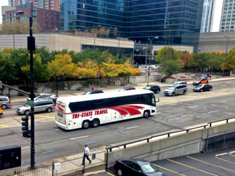 Street scene with a white bus, cars, and a few pedestrians near a city building.