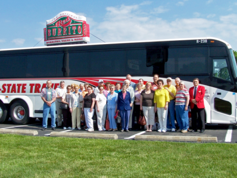 Group posing beside a large black and red tour bus in front of a state travel sign on a sunny day