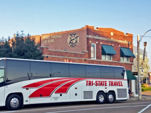 Tri-State Travel coach bus parked beside a brick building with a mural and green awnings