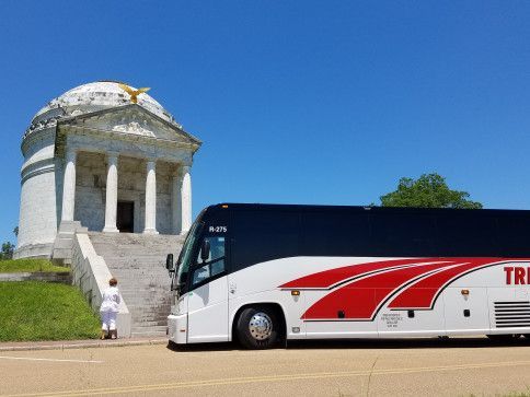 White tour bus parked beside a stone memorial under a clear blue sky