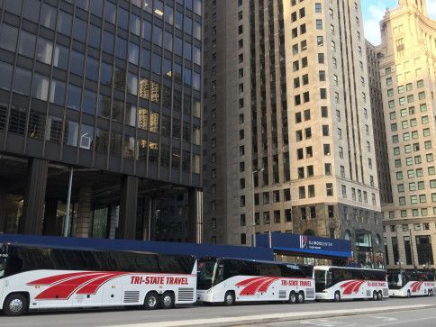 Five white-and-red buses lined up on a city street beside tall office buildings.