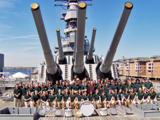 Large group posing on a battleship deck in front of huge naval guns under a blue sky