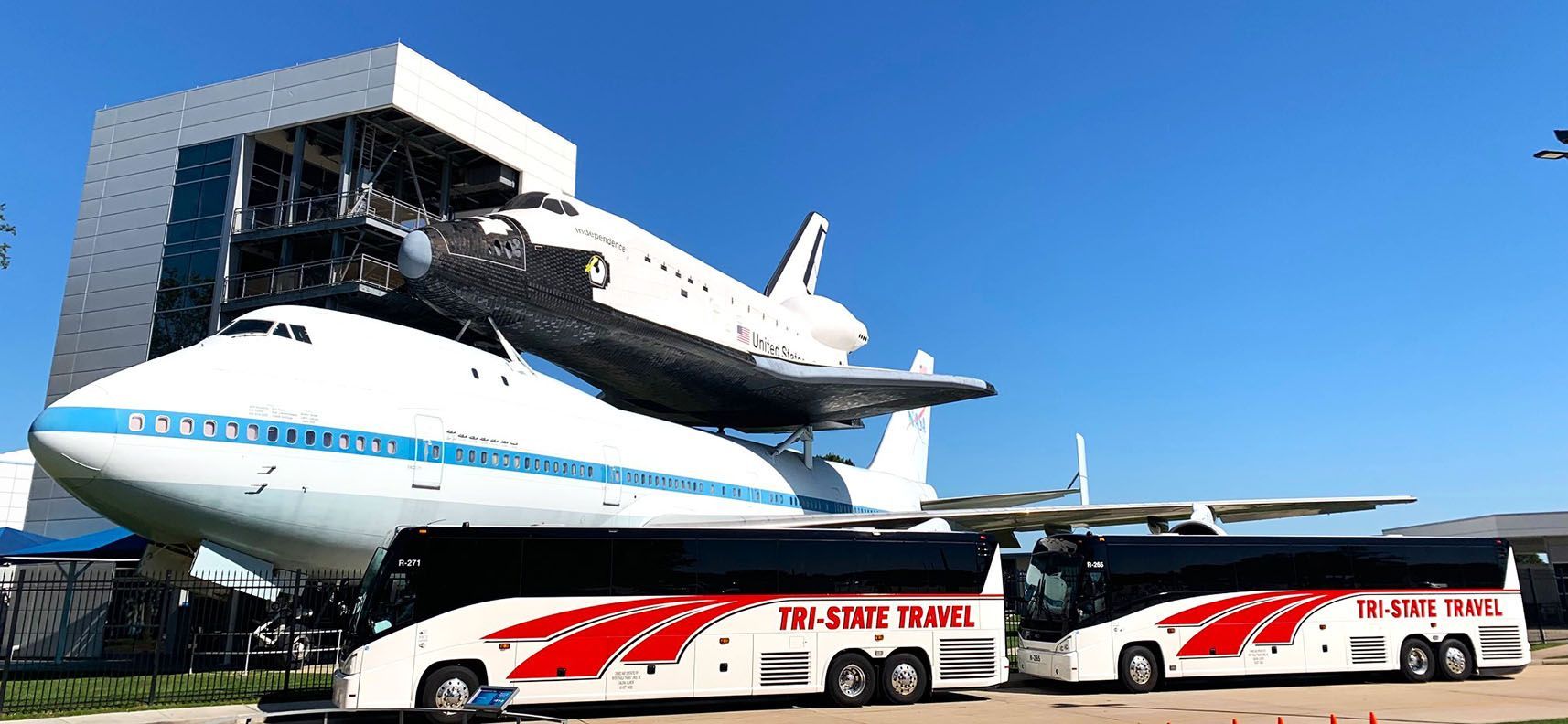 Space shuttle mounted on a Boeing 747 with two white-and-red buses below in front of a modern building.