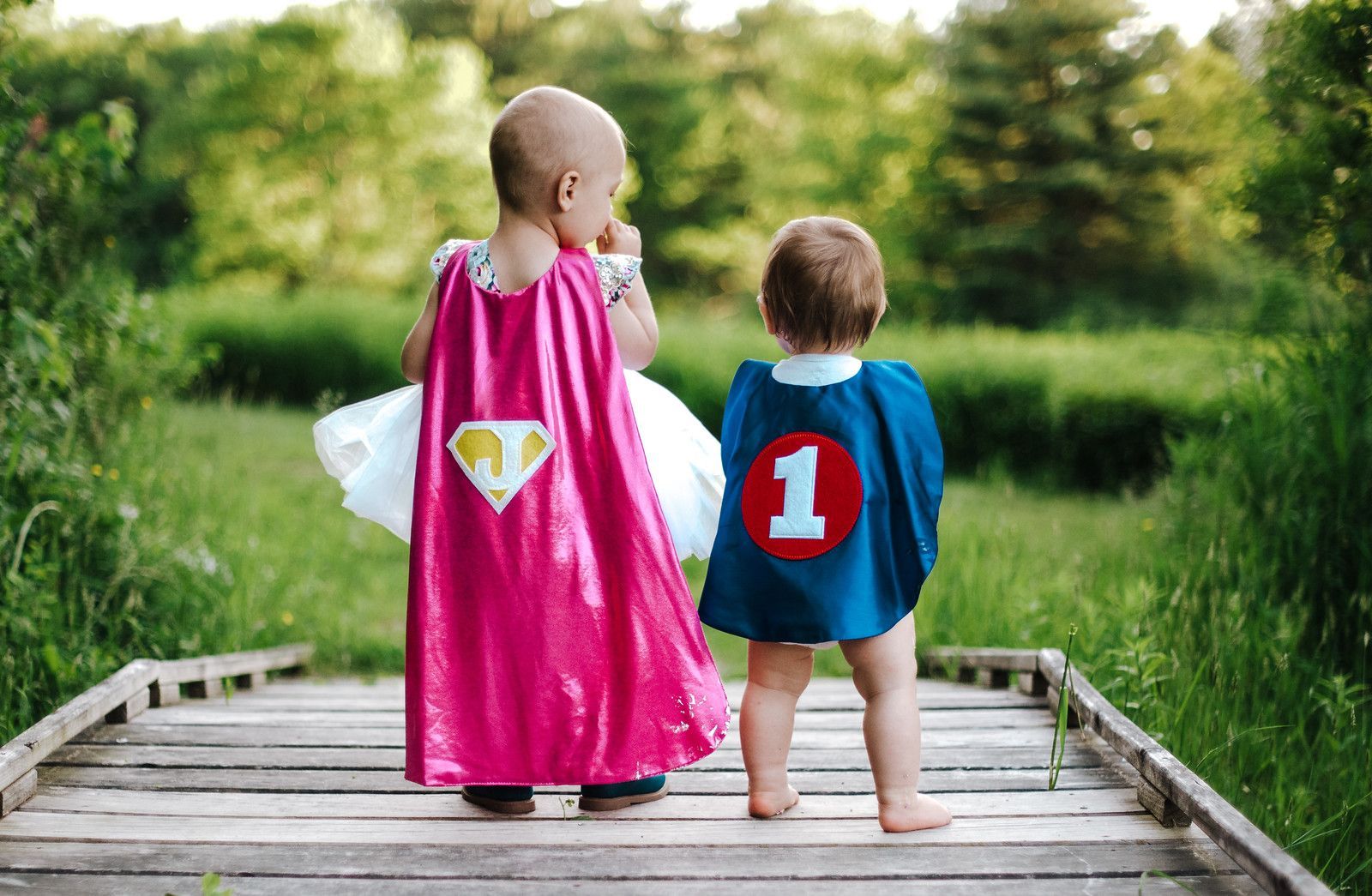 Two children in superhero capes standing on a wooden bridge in a green park