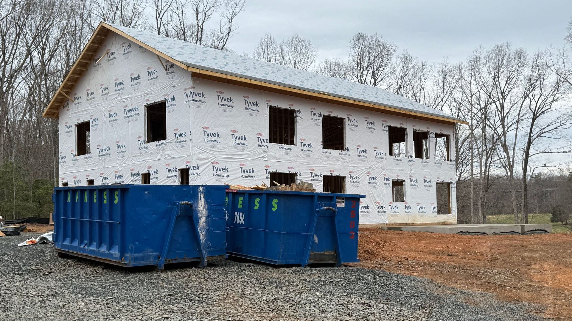 Construction site of a two-story building wrapped in white protective material; blue dumpsters sit in the foreground.
