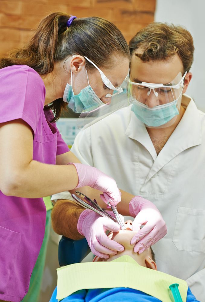 Dentist and assistant treating a patient. They wear masks, gloves, and safety glasses. Pink scrubs are visible.