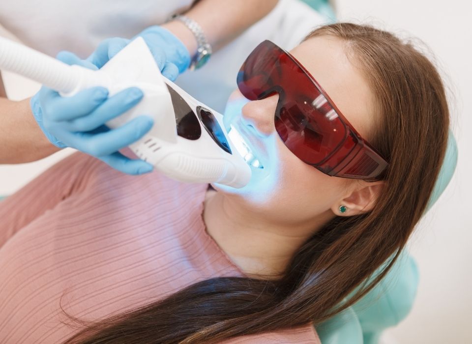 Woman in dental chair receiving teeth whitening treatment, wearing protective eyewear.