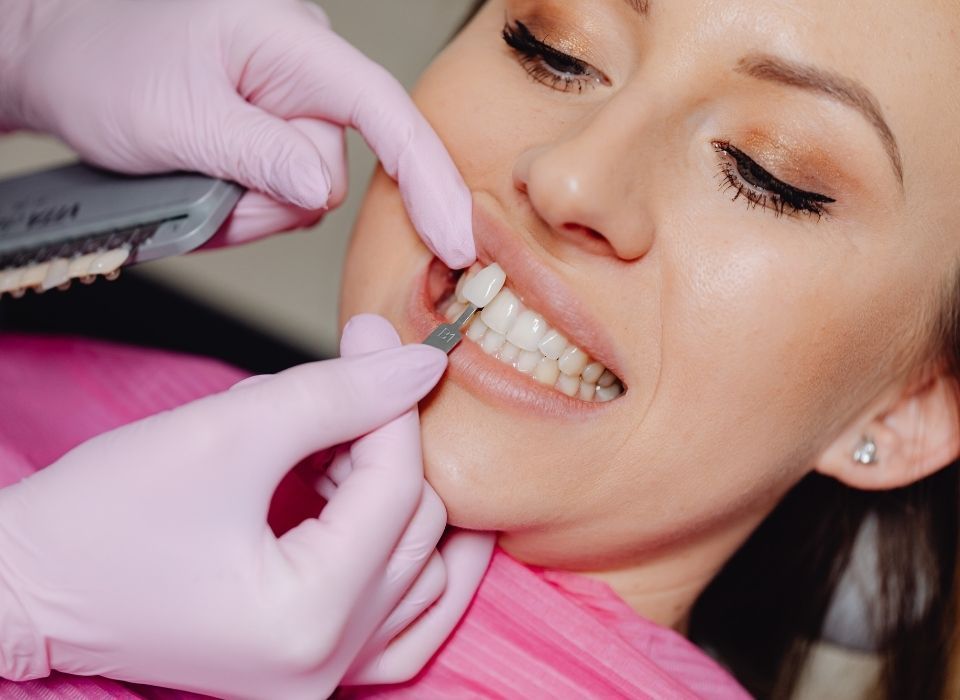 Dentist comparing tooth shade with a guide for a patient in a dental office, wearing pink gloves and shirt.