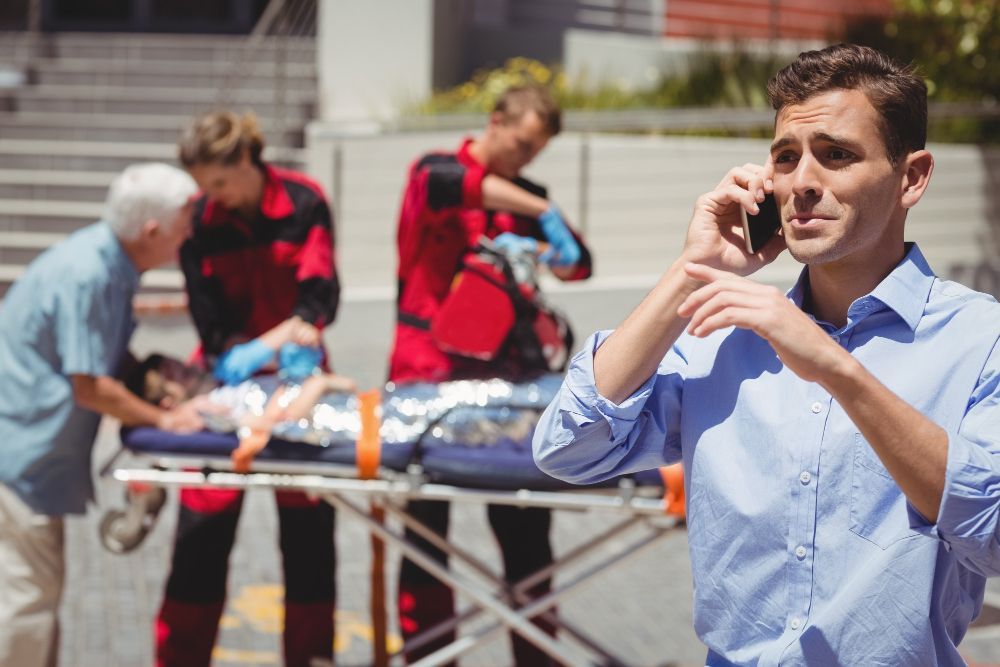 El hombre al teléfono mientras el personal médico de emergencia atiende a una persona en camilla al aire libre.