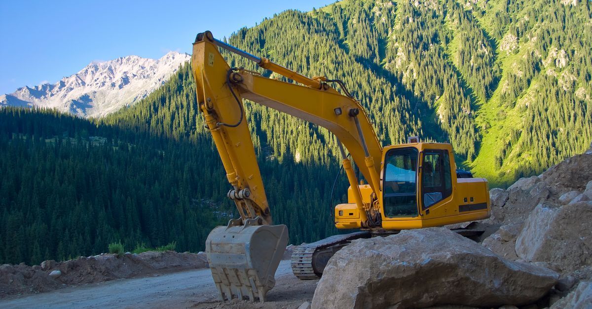 Yellow excavator on a dirt site with forested mountains in the background