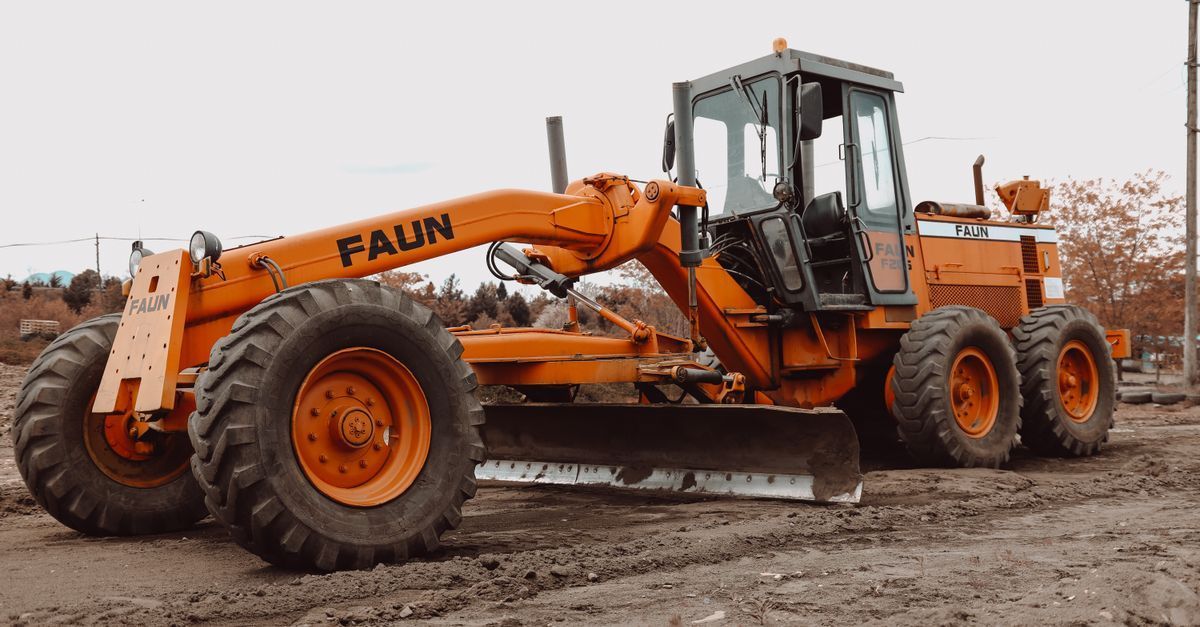 Orange road grader parked on muddy ground, with large black tires and enclosed cab