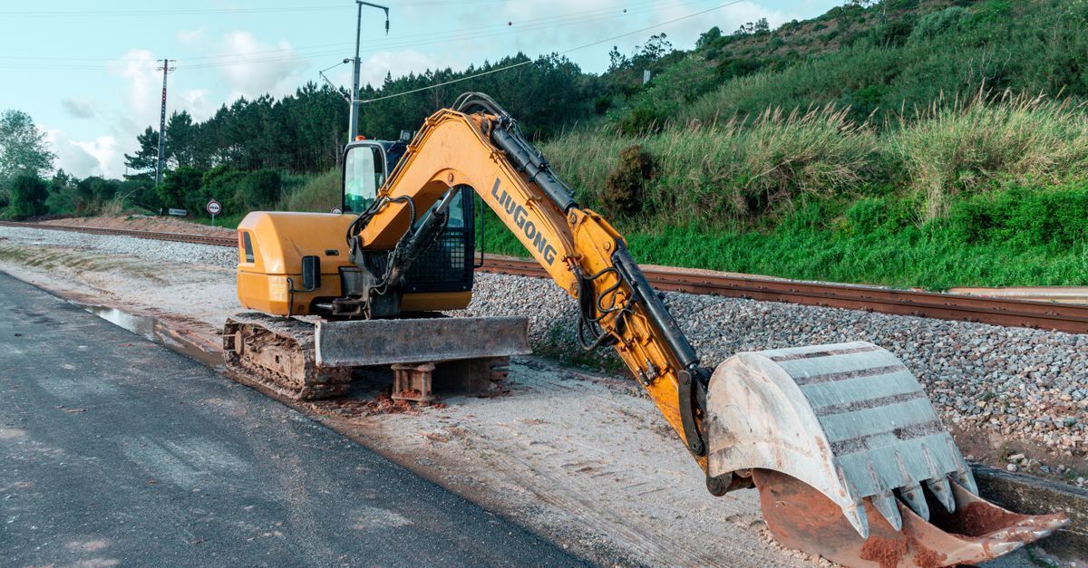 Yellow excavator on a roadside, digging dirt beside a green embankment.