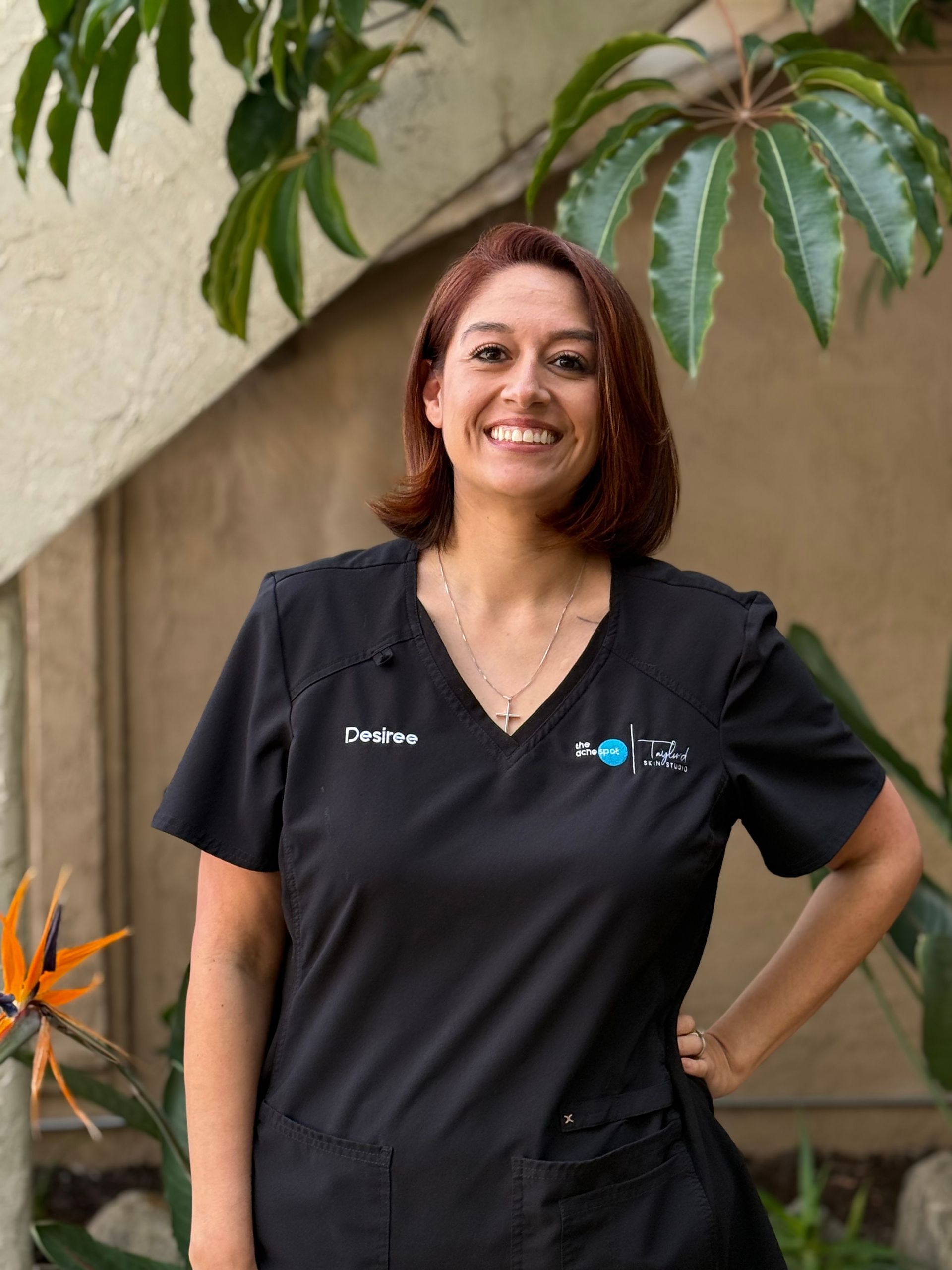 Woman with glasses smiles, wearing black scrubs, standing outdoors near green leaves.