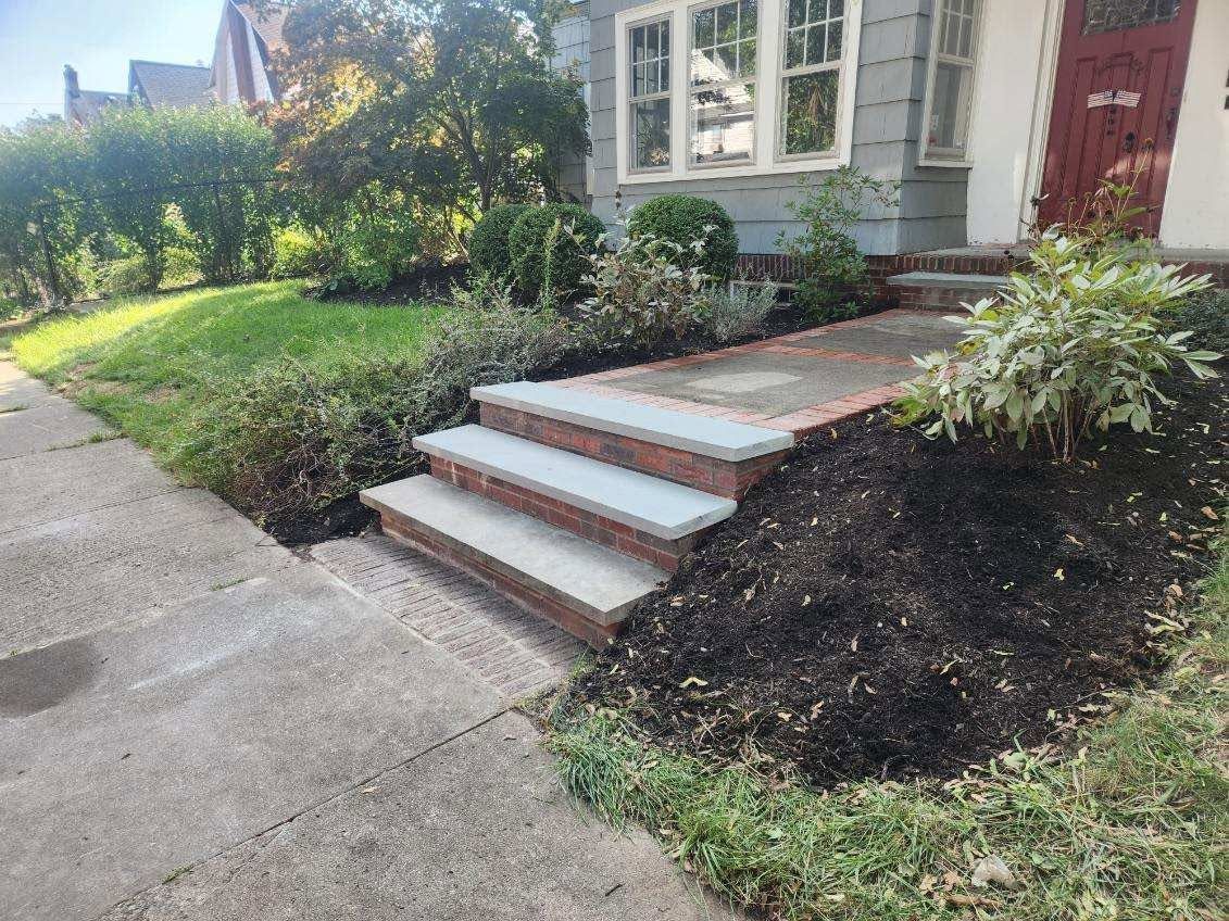 Front steps to a house with white trim, red door, and landscaped mulch beds beside a concrete walkway