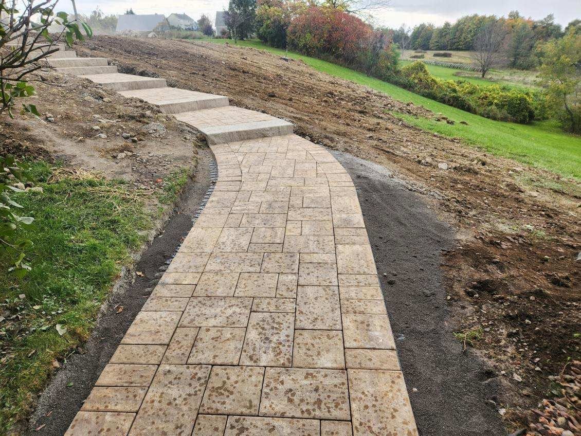 Curved stone walkway under construction beside grassy fields and dirt embankments