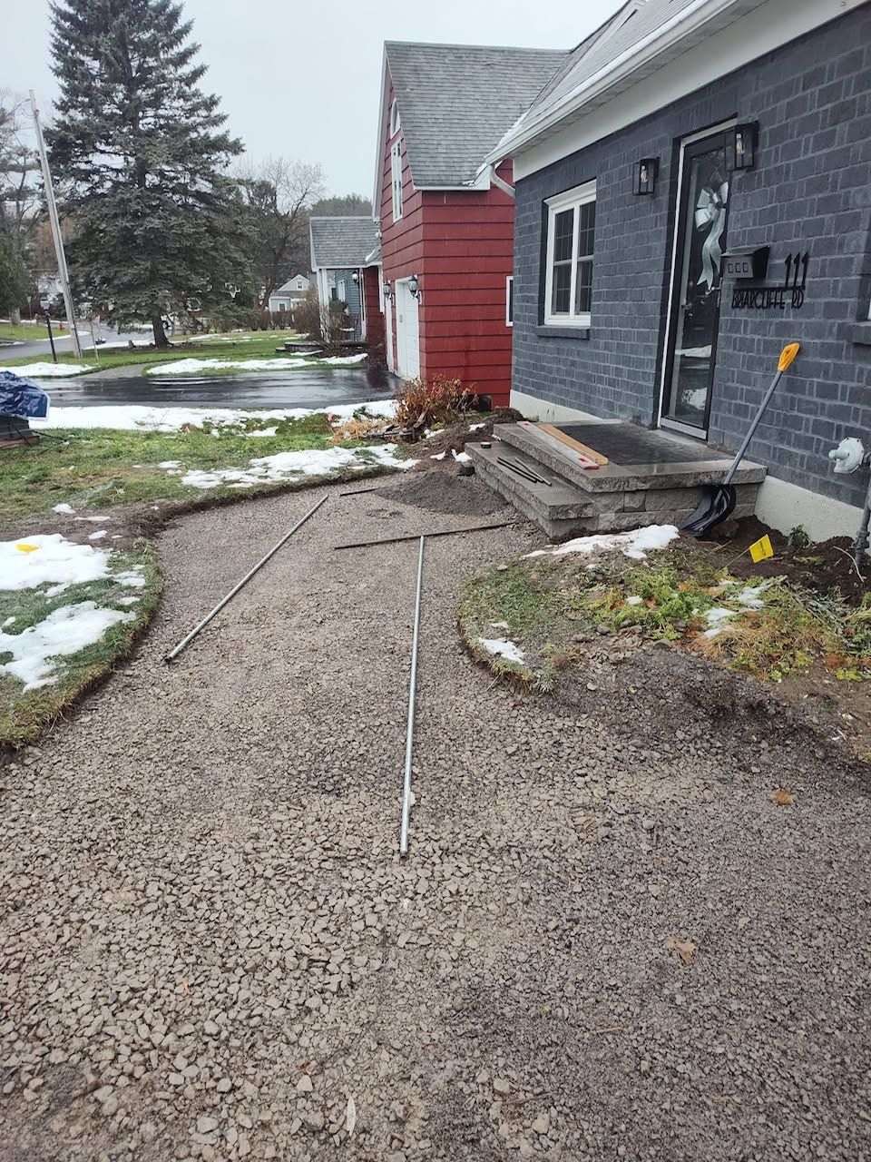 Rough gravel walkway beside a gray house with snow patches, a red building, and fallen debris after storm damage
