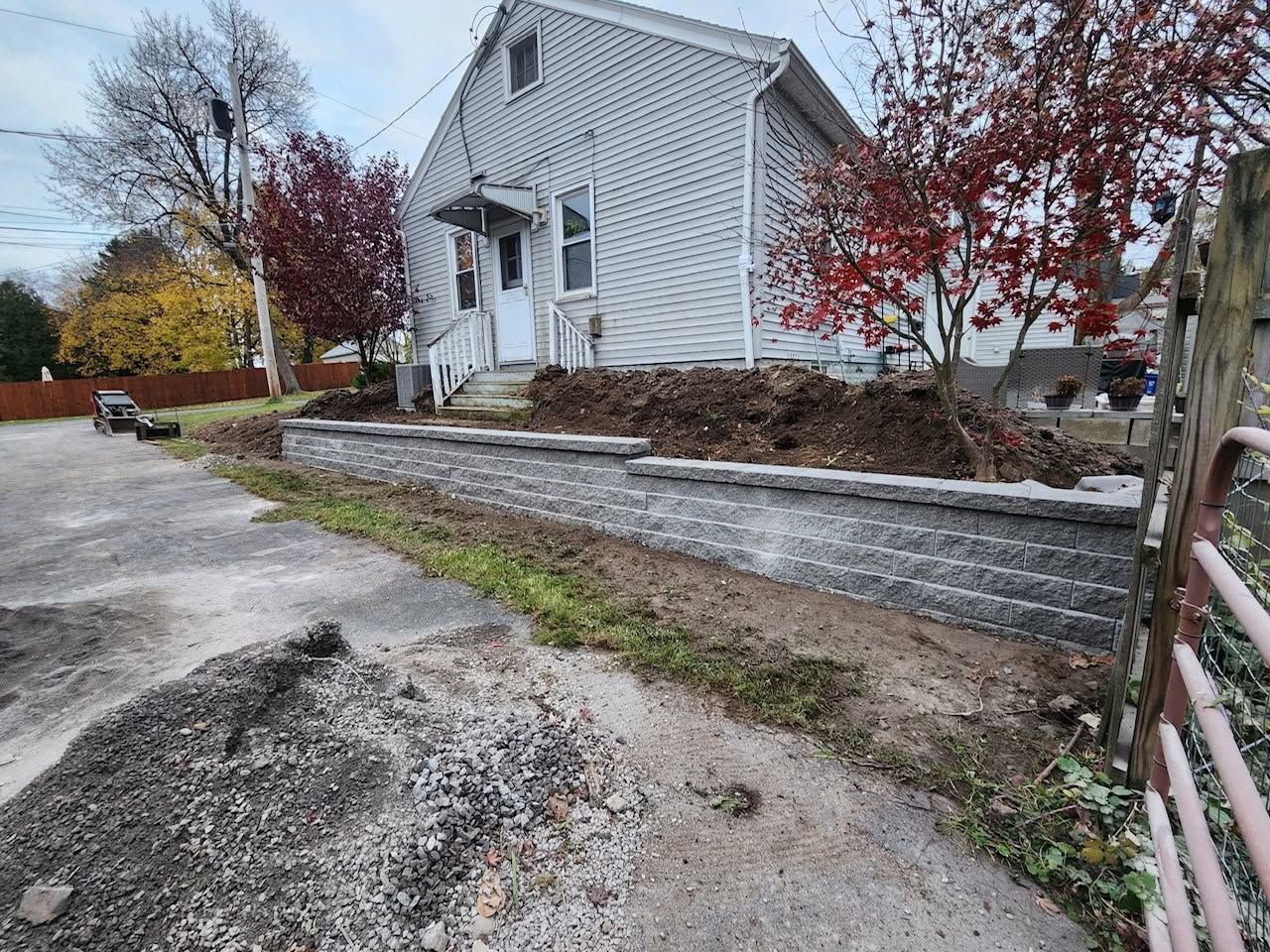 White house by a gravel driveway with stone retaining wall and autumn trees beyond a metal gate