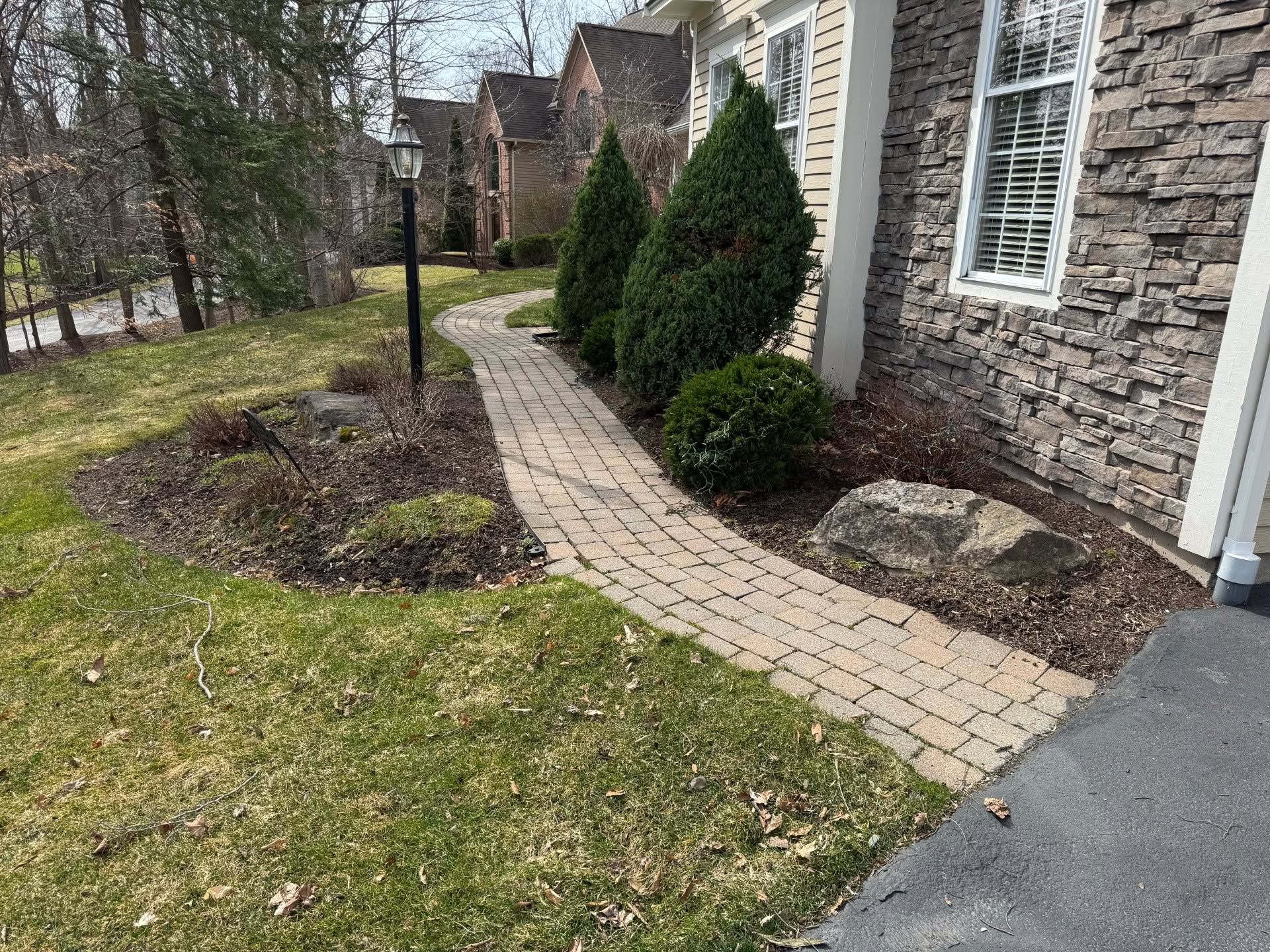 Curved brick walkway beside a stone house, with trimmed shrubs and a lawn in the foreground.