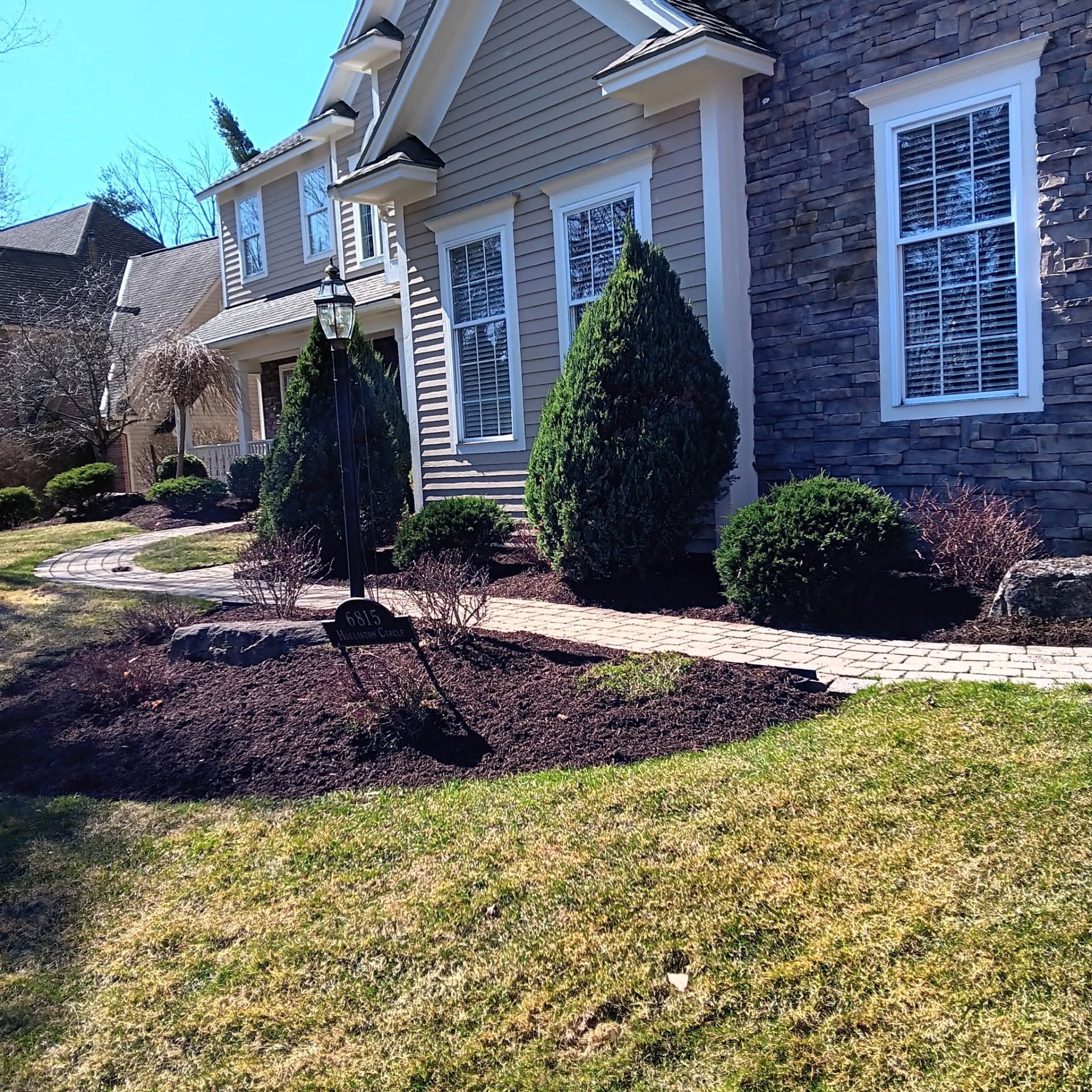 Front yard of a suburban house with stone siding, white-trimmed windows, shrubs, and a curving walkway.
