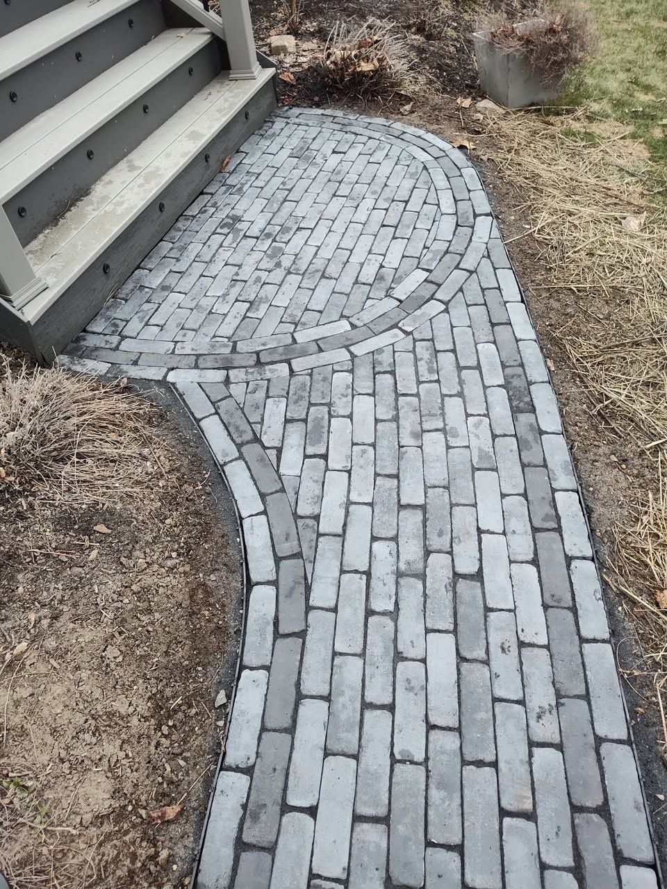 Curved gray brick walkway beside house steps, bordered by dry grass and landscaping stones