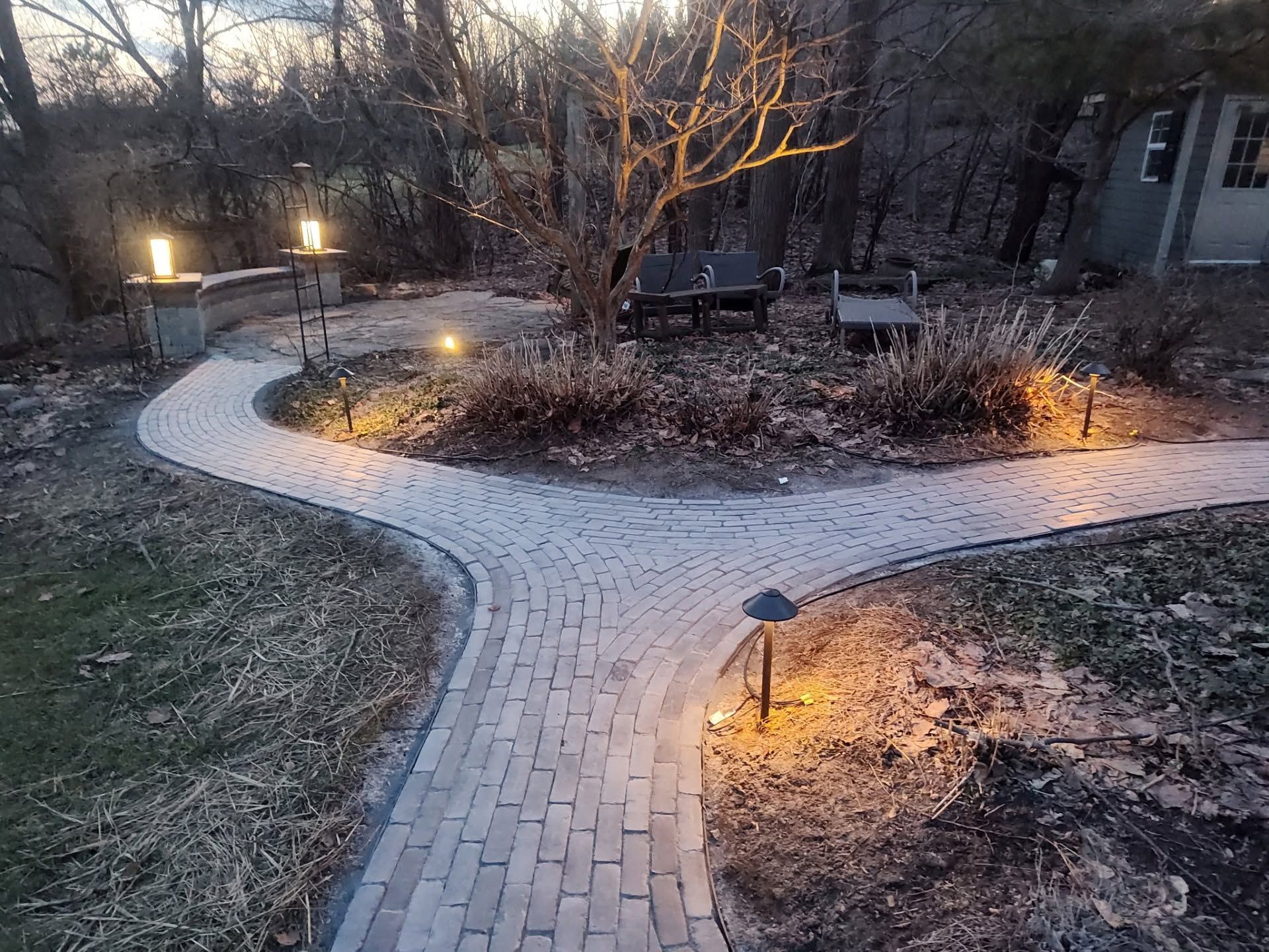 Curved brick garden path at dusk, lit by small landscape lights around bare shrubs and a house.