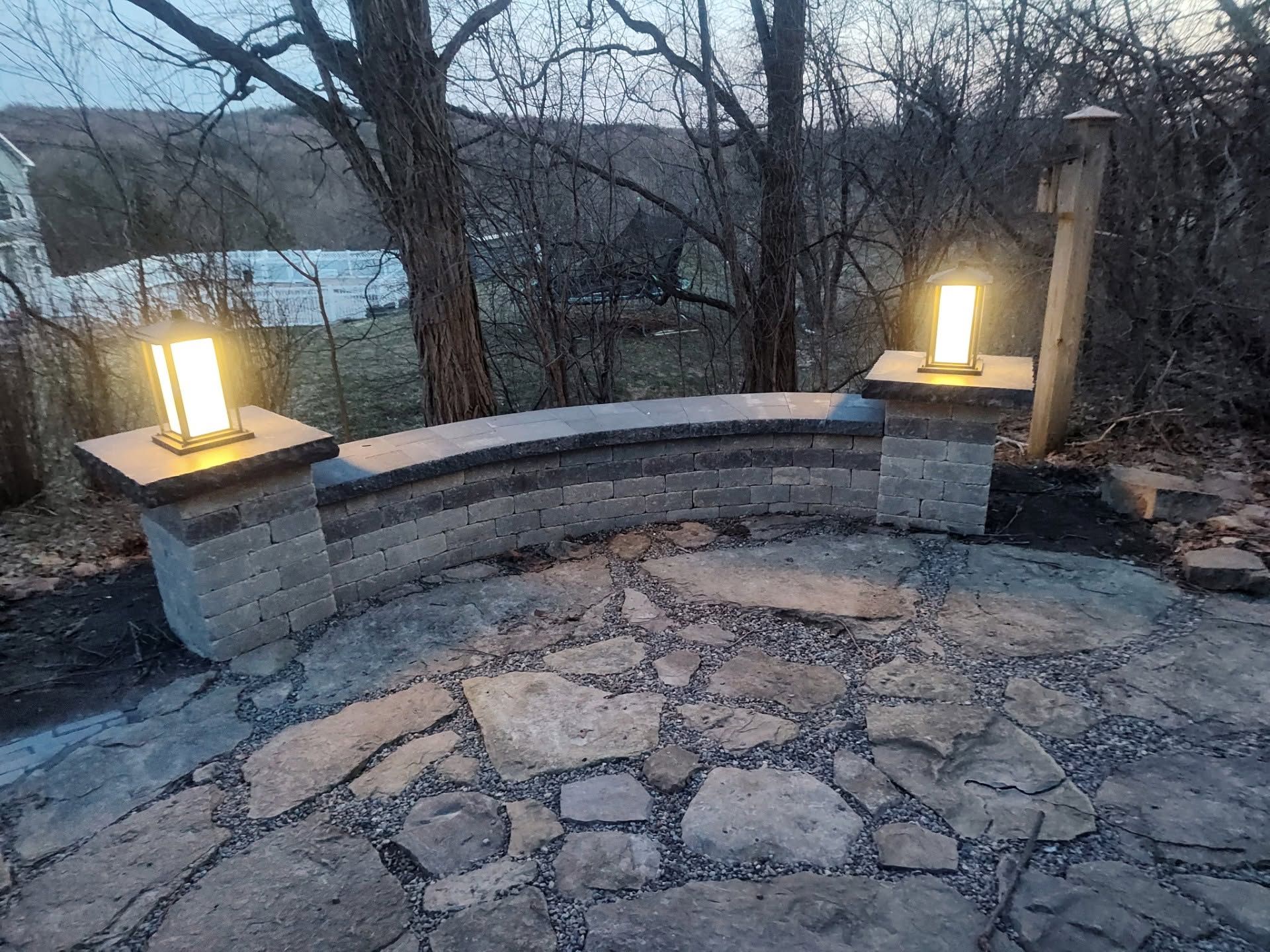 Stone patio with curved bench and two lit lanterns at dusk overlooking trees and a lake