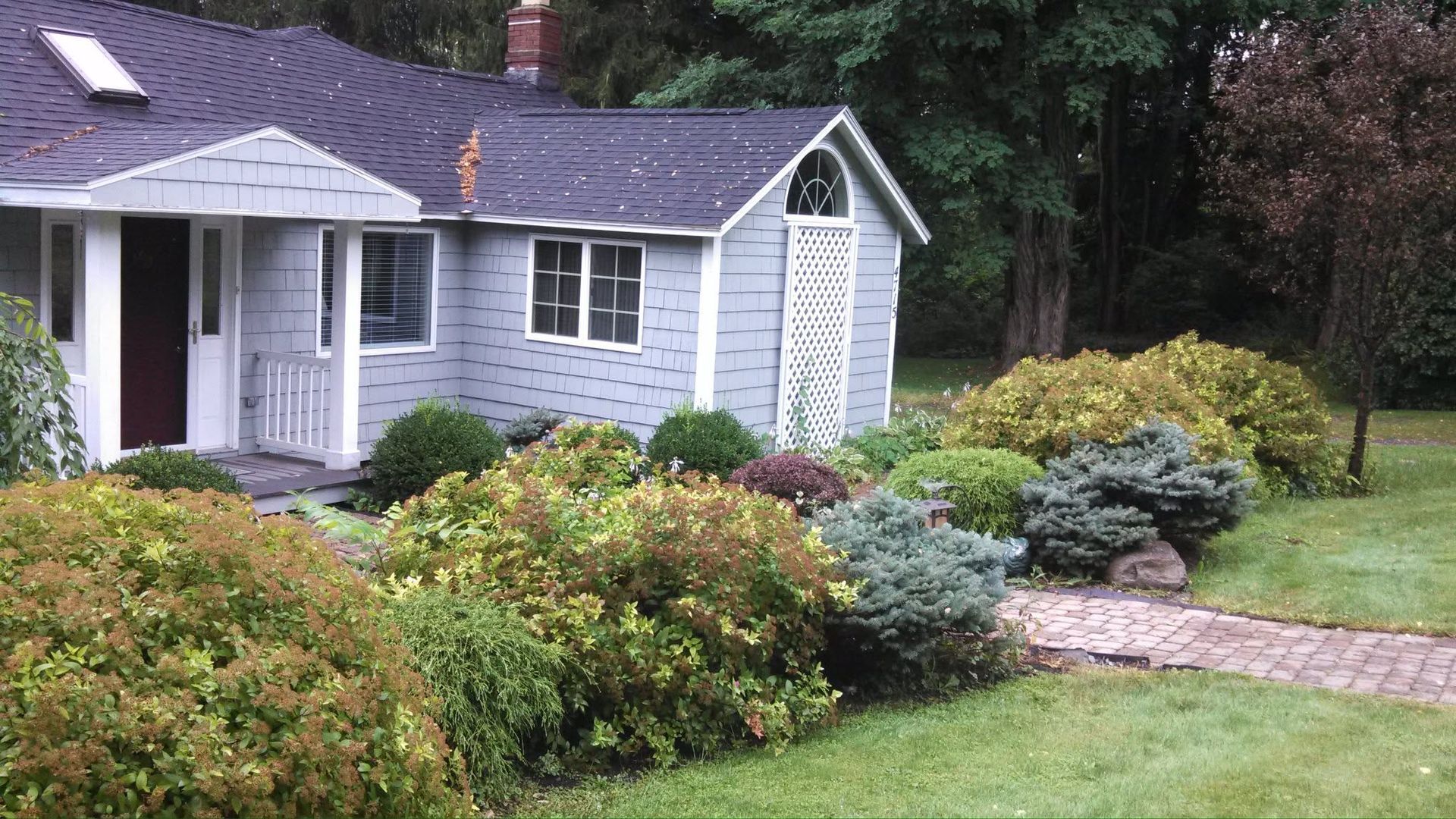 Small gray house with white trim, surrounded by lush shrubs and a brick path in a green yard
