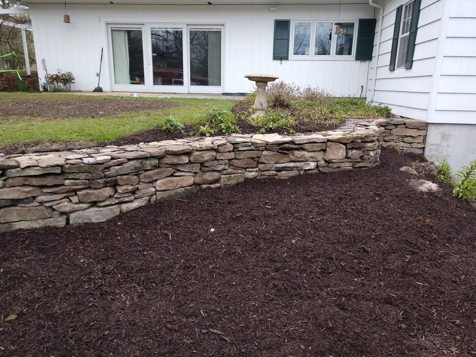 Stone retaining wall with mulch bed and lawn in front of a white house with sliding glass doors