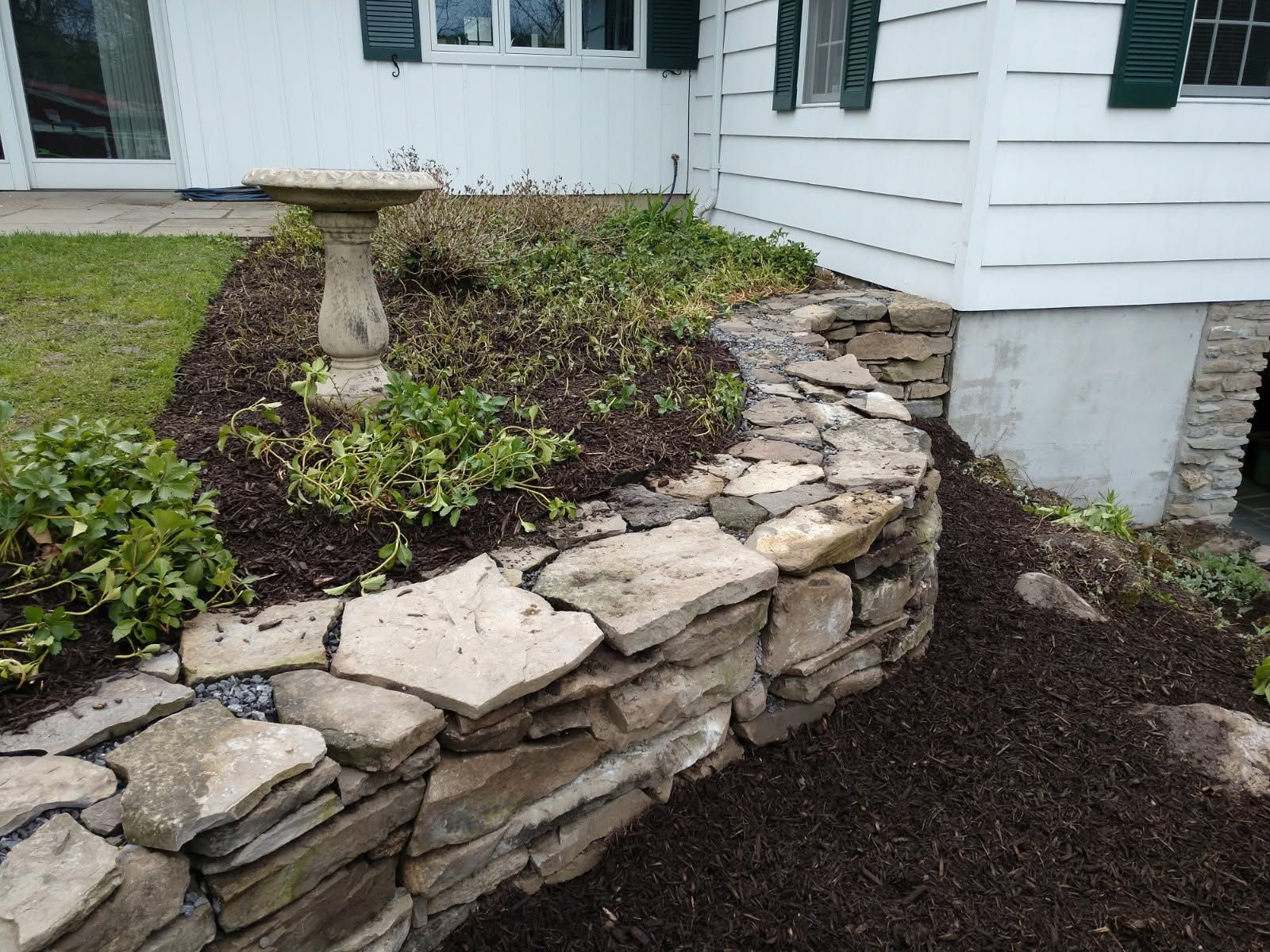 Stone retaining wall with dark mulch and a birdbath beside a white house porch