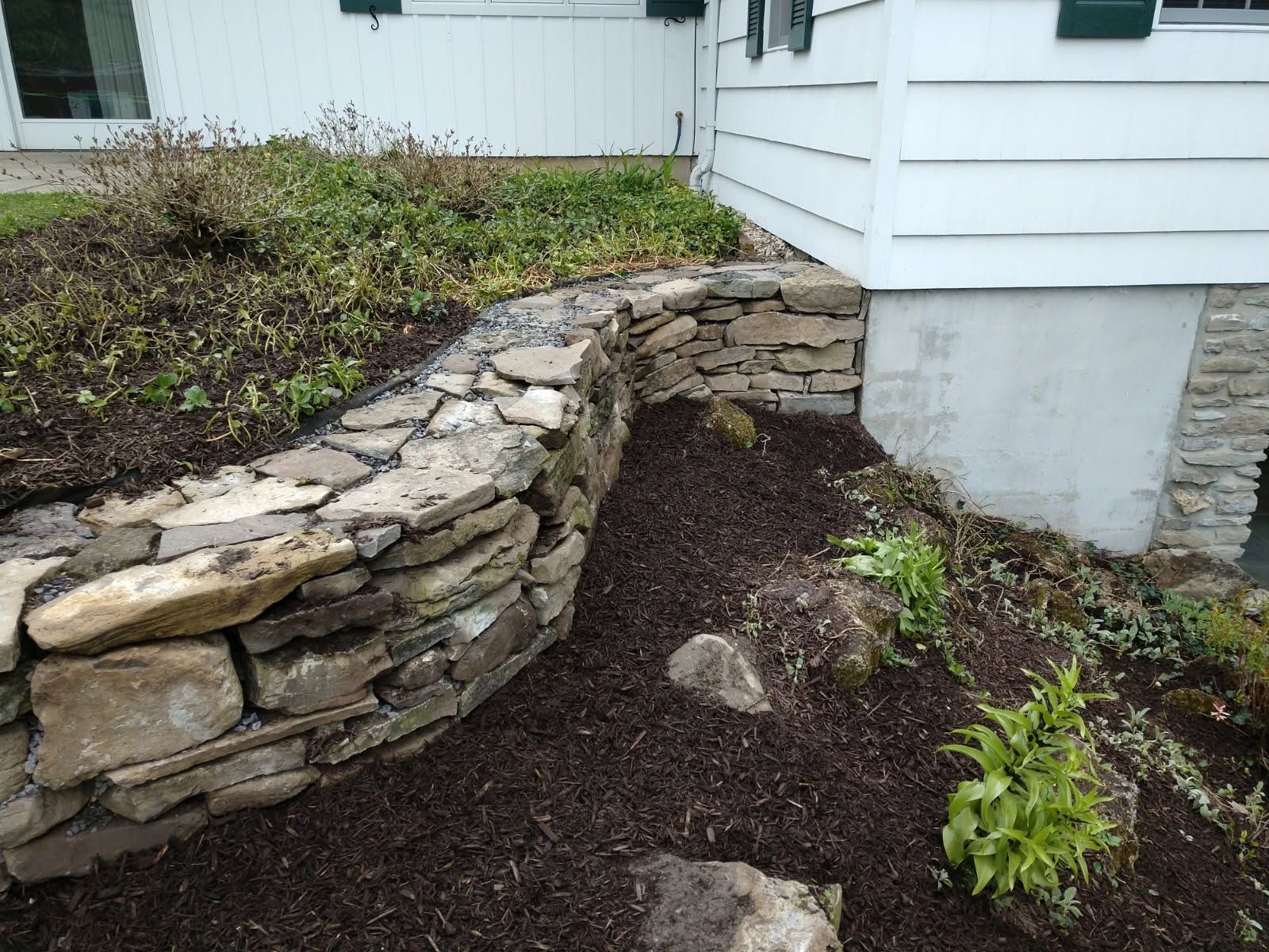 Stone retaining wall beside a white house, with dark mulch and a small green plant below.