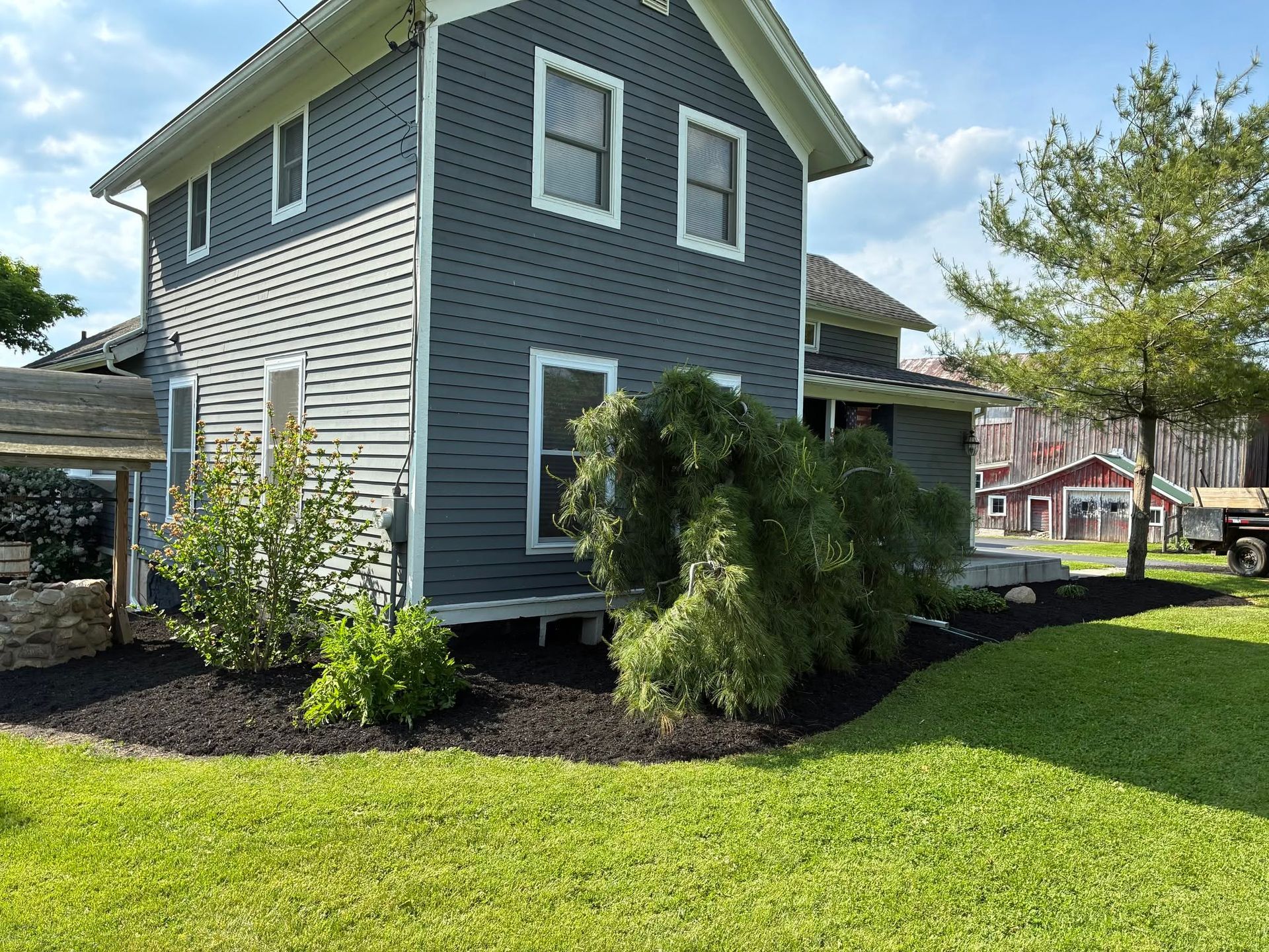 Blue-gray house with white trim, green lawn, and shrubs under a partly cloudy sky