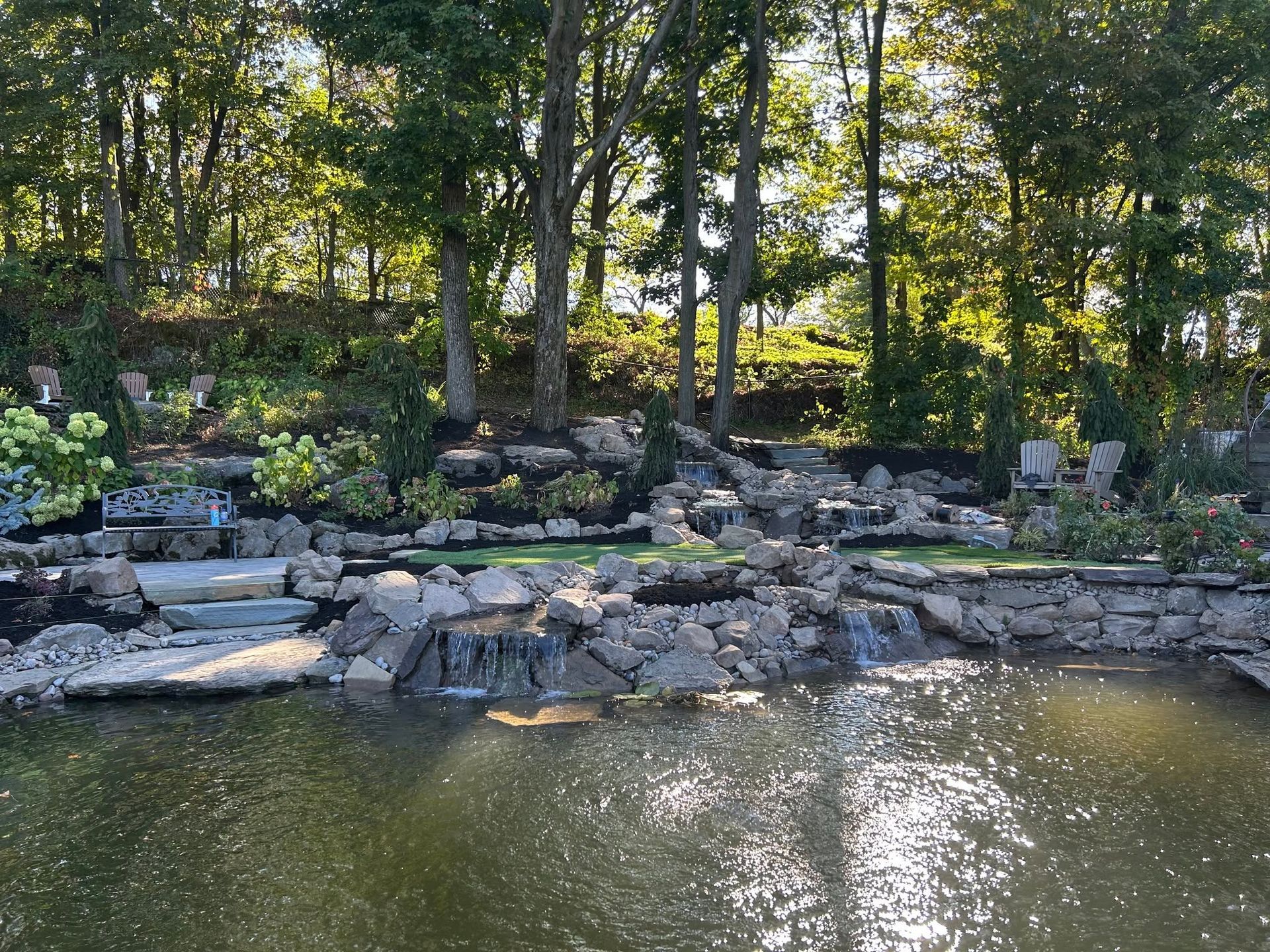 Stone patio with wooden benches and Adirondack chairs in a green garden setting