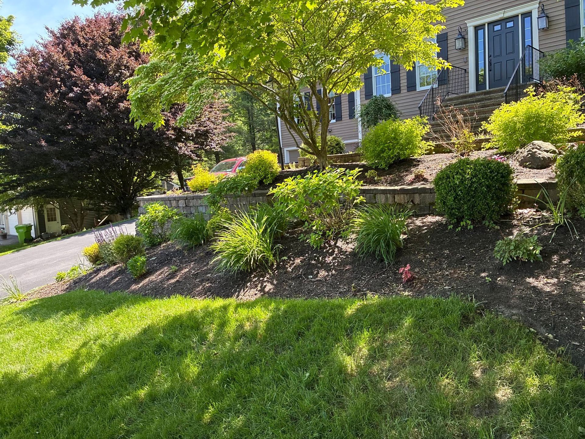 Front yard garden with stone retaining wall, green lawn, flowering shrubs, and a house porch in sunlight